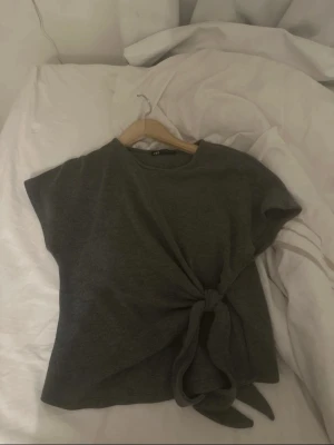 Mörkgrå topp med knyt från Zara - Snygg mörkgrå topp från Zara med rund hals och korta ärmar. Modellen har ett coolt knyt framtill som ger en extra touch. Mjukt material och enkel, rak passform. Perfekt att styla med jeans eller kjol för en chill look.