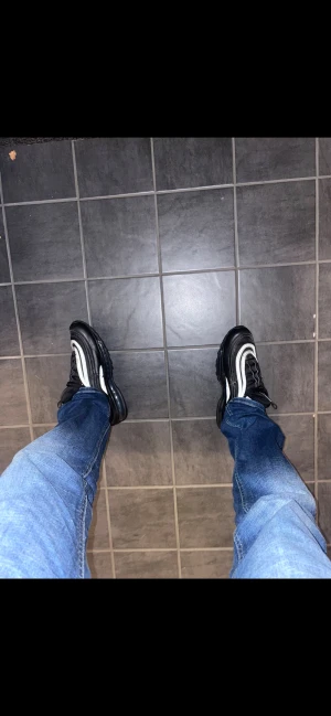 Nike Air Max 97 svart och vit - Nike Air Max 97 sneakers i svart och vit med ikoniska vågiga linjer och synlig Air-sula. Ovandel i syntet och mesh, snörning och rund tå. Snygg design med reflekterande detaljer och diskret blå Nike-logga på sidan och tungan.