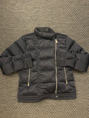 Svart pufferjacka från Boomerang - Svart pufferjacka från Boomerang med hög krage, guldiga dragkedjor och snygga detaljer vid ärmsluten. Jackan har quiltad design och två dragkedjeförsedda fickor framtill. Perfekt för kalla dagar och ger en cool vibe till din outfit.