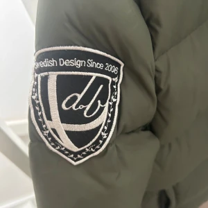 Olivgrön pufferjacka från db - Snygg olivgrön pufferjacka från db med broderad patch på ärmen, huva och dragkedja framtill. Jackan har flera fickor med dragkedja och en cool, quiltad look. Perfekt för kalla dagar när du vill hålla stilen.