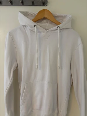 Vit hoodie från Pier One med huva - Vit hoodie från Pier One med klassisk huva och snörning. Tröjan har en stor magficka framtill och är långärmad. Tillverkad i mjuk bomull, perfekt för chill dagar. Enkel och stilren design utan mönster eller detaljer.