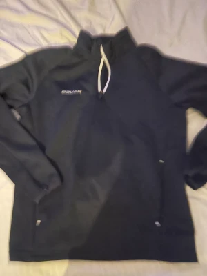 Svart Bauer half zip tröja - Svart långärmad tröja från Bauer med half zip och vit snörning i halsen. Perfekt för träning eller chill, med diskret Bauer-logga på bröstet. Skön passform och sportig look.