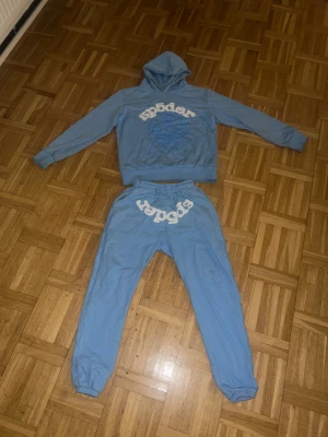 Full blå tracksuit set från sp5dee - Säljer ett par blå mjukisbyxor från Sp5der med vit logga tryckt över rumpan. Byxorna har resår i midjan och vid bensluten för skön passform. Materialet är mjukt bomullstyg, perfekt för chill dagar. Matcha med hoodie för en komplett look.