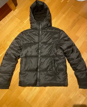 Svart pufferjacka från Studio Total - Svart pufferjacka från Studio Total i storlek S med huva och dragkedja framtill. Jackan har en clean look med två fickor på sidorna och är vadderad för extra värme. Perfekt för kalla dagar och enkel att matcha med allt.
