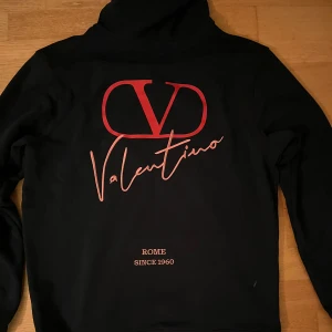 Mörkblå Valentino hoodie med röda och rosa detaljer  - Mörkblå hoodie från Valentino med stor röd logga och text på ryggen samt mindre logotyp på bröstet. Tröjan har huva med snörning och en klassisk magficka. Materialet är mjukt och passar perfekt för dig som gillar exklusiv streetwear. Lappen inuti tröjan är halvt av, men sitter kvar på. Fråga gärna om bild. Icke org!nal