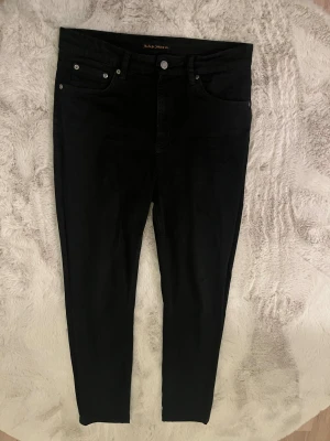 Svarta skinny jeans från Nudie - Snygga svarta jeans från Nudie Jeans Co med klassisk femficksdesign och silverfärgade nitar. Modellen har smal passform och raka ben, med Nudie-logga på bakfickan och läderpatch i midjan. Perfekta för dig som gillar stilrena och enkla jeans.