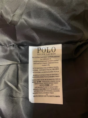 Svart pufferjacka från Polo Ralph Lauren - Svart pufferjacka från Polo Ralph Lauren med röd broderad logga på bröstet. Jackan har dragkedja framtill, två sidofickor med dragkedja och huva. Materialet är bomull, vilket ger en lätt och sportig känsla. Perfekt för kalla dagar.