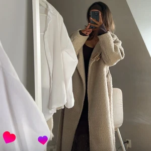 Beige teddykappka med bälte från Gina Tricot - Säljer en beige teddykappa från Gina Tricot med bred krage och bälte i midjan. Jackan är lång, har en loose passform och är tillverkad i mjukt fleece-liknande material. Perfekt för dig som gillar oversized och mysiga vibes. Har använt min under vintern och den håller dig extremt varm! 