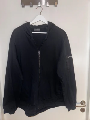 Svart hoodiejacka från Emporio Armani - Svart hoodiejacka från Emporio Armani med dragkedja framtill och huva med snörning. Jackan har lång ärm, diskret logga på ärmen och är tillverkad i mjuk bomull. Perfekt för en chill och stilren look.