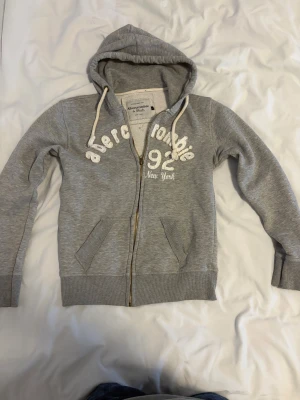 Grå hoodie Abercrombie & Fitch - Snygg grå hoodie från Abercrombie & Fitch med vit broderad logga och siffror på bröstet. Tröjan har huva med snörning, dragkedja i guld och två fickor framtill. Perfekt för chill dagar och streetstyle.