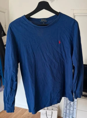 Blå långärmad tröja från Polo Ralph Lauren - Snygg marinblå långärmad tröja från Polo Ralph Lauren med klassisk rund hals och röd broderad logga på bröstet. Tillverkad i mjuk bomull, perfekt för chill dagar. Enkel och stilren design som funkar till jeans eller shorts.