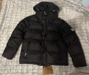 Svart pufferjacka från Burberry - Snygg svart pufferjacka från Burberry med huva och tydlig logga på ärmen. Jackan har dragkedja framtill, två fickor med dragkedja och quiltad design. Perfekt för kalla dagar och ger en clean och modern look.