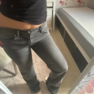 Grå bootcut jeans med låg midja - Snygga grå jeans med bootcut och låg midja. Klassisk femficksmodell med silverfärgade nitar och knappar. Jeansen har en rak passform över låren och lite utsvängda ben. Obs byxorna har slitage längst ner
