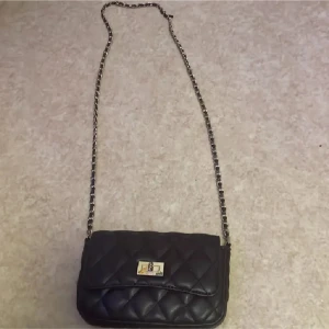 Brun quiltad axelväska med kedja - Stilren brun axelväska med quiltat mönster och lång kedjerem i guld och svart. Väskan har ett snyggt guldfärgat lås framtill och ett rymligt fack med dragkedja inuti. Perfekt storlek för det viktigaste och ger en cool vibe till din outfit.