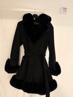 Svart pälsjacka med bälte - Snygg svart pälsjacka med stora pälskanter vid krage, ärmslut och nederkant. Jackan har ett bälte i midjan för extra stilpoäng och markerad siluett. Perfekt för dig som vill sticka ut med en lyxig och trendig look.