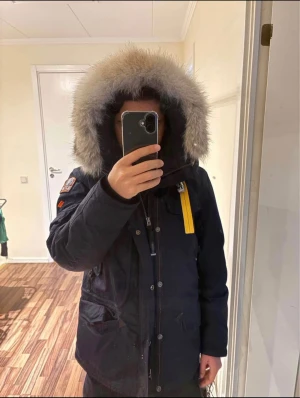 Parajumpers svart dunjacka med päls - Mörk blå dunjacka från Parajumpers med stor huva och fluffig päls runt kanten. Jackan har en gul rem vid halsen, flera fickor och patch på ärmen. Långärmad och med både dragkedja och knappar framtill. Perfekt för kalla dagar och riktigt snygg streetstil. Köpt för: 11 000kr Storlek: Small Använd 3 gånger. Går ner vid snabb affär. 