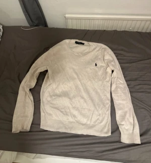 Beige tröja från Polo Ralph Lauren - Snygg beige tröja från Polo Ralph Lauren i mjuk pima bomull. Klassisk rund hals och långärmad modell med den ikoniska svarta Polo-loggan broderad på bröstet. Perfekt för dig som gillar stilrena och tidlösa plagg.