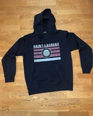 Svart YSL Hoodie - Svart hoodie frän Saint Laurent Paris med tryck i vitt och rött framtill, använd fätal gänger. Klassisk känguruficka, snörning i huvan och ribbade muddar. Perfekt för dig som gillar streetstyle och vill ha nagot snyggt och bekvämt. Skriv privat för mer information.