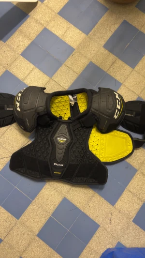CCM Tacks 6052 SR M - CCM Tacks 6052 axelskydd i storlek Medium Senior. Svart och gul design, robust skydd för hockey. Synligt märke och modell, lättare yttre slitage men i gott skick. Perfekt för spelare som söker pålitligt skydd och komfort.