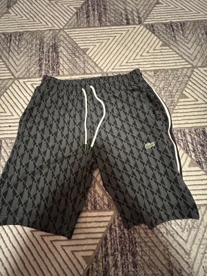 Svarta mönstrade shorts från Lacoste - Snygga svarta shorts från Lacoste med grått geometriskt mönster och klassisk krokodillogga. De har vit snörning i midjan och vita ränder längs sidorna. Perfekta för chill dagar och sportiga outfits. Materialet är mjukt och känns som polyester.
