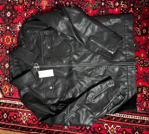 Svart skinnjacka med dragkedjor - Snygg svart skinnjacka med raka linjer och coola detaljer som dragkedjor vid ärmsluten och bröstfickor med knappar. Jackan har klassisk krage och stängs med dragkedja framtill. Perfekt för dig som vill ha en stilren och edgy look.