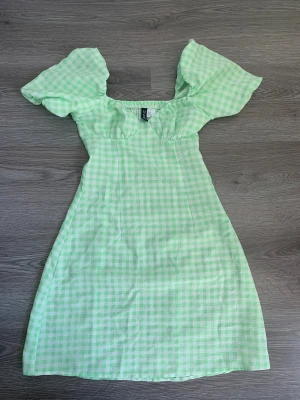 Grön rutig klänning från H&M  - Superfin ljusgrön och vit rutig klänning från H&M i storlek XS. Klänningen har puffiga korta ärmar och en söt, figurnära passform. Perfekt för vår och sommar med sitt lätta bomullsmaterial och fräscha färg.
