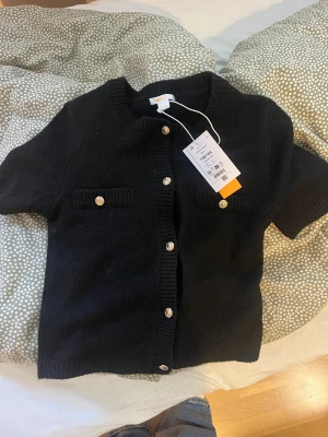 Svart stickad kofta med kort ärm - Svart stickad kofta från Gina Tricot med korta ärmar och snygga guldfärgade knappar framtill. Koftan har två dekorativa fickor på bröstet och ribbade kanter vid hals, ärmslut och nederkant. Perfekt för dig som gillar klassisk och stilren look.