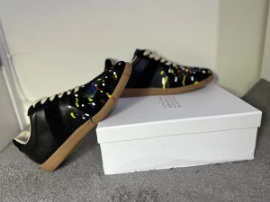 Maison Margiela Gats sneakers med färgstänk - Svarta sneakers från Maison Margiela med coolt färgstänksmönster i gult, vitt, rött och blått. Skorna har beige snörning, rund tå och platt gummisula i brunt. Tillverkade i skinn och mocka för en lyxig känsla. Perfekt för dig som vill sticka ut med din stil.