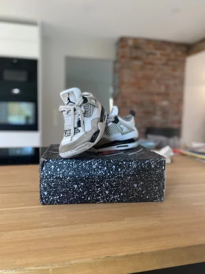 Nike Air Jordan 4 Retro vit/svart - Nike Air Jordan 4 Retro sneakers i vit och svart med beige detaljer. Skorna har snörning, meshpaneler och platt sula med synlig Air-enhet. Ikonisk Jumpman-logga på tungan och coola plastdetaljer på sidorna. Perfekt för dig som gillar streetstyle och klassiska sneakers.