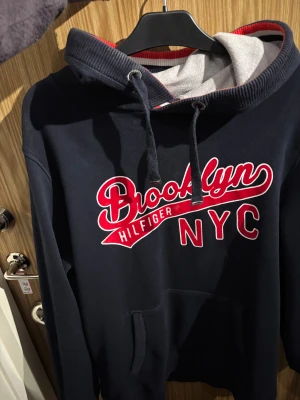 Mörkblå Hilfiger hoodie Brooklyn NYC - Snygg mörkblå hoodie från en Hilfiger Denim med stor Brooklyn NYC-text i rött och vitt framtill. Tröjan har huva med snörning, känguruficka och mjukt grått foder i luvan. Perfekt för dig som gillar streetstyle och amerikansk vibe.