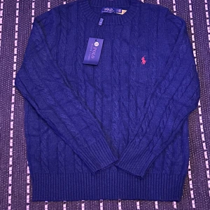 Marinblå stickad tröja Polo Ralph Lauren - Snygg marinblå stickad tröja från Polo Ralph Lauren med klassiskt kabelstickat mönster och röd broderad logga på bröstet. Rund hals och ribbade muddar vid ärmslut och nederkant. Perfekt för dig som gillar stilrena och tidlösa plagg. Priset kan prutas