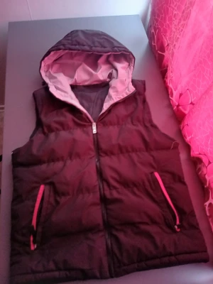 Svart dunväst med huva och dragkedja - Svart dunväst med grå insida på huvan och snygga röda detaljer vid fickorna. Västen har dragkedja framtill och två fickor med röda dragkedjor. Perfekt för lager-på-lager och chill stil under höst och vinter.