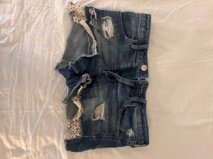 Blå jeansshorts med spetsdetaljer - Snygga blå jeansshorts från Bik Bok med slitna detaljer och råa kanter. Shortsen har vit spets längs sidorna och flera coola hål och slitningar framtill. Klassisk femficksmodell med knapp och dragkedja. Perfekta för sommarens alla äventyr!