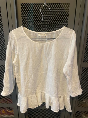 Vit blus i linne med volangkant - Supersöt vit blus från Vintage i luftigt linne. Blusen har trekvartsärmar, rund halsringning och en volangkant längst ner som ger en romantisk vibe. Perfekt för dig som gillar en enkel men ändå detaljrik stil.
