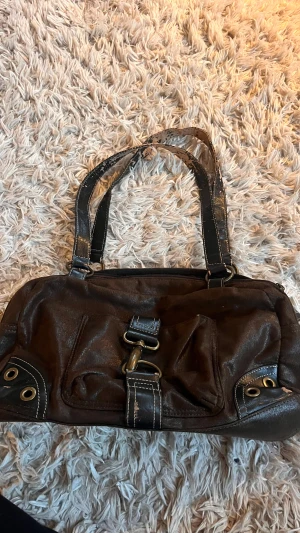 Brun axelväska från Martti - Snygg brun axelväska från Martti i skinnimitation med coola metalldetaljer och kontrastsömmar. Väskan har praktiska fack med dragkedja både fram och bak, samt ringar och öglor som ger en edgy vibe. Perfekt storlek för allt du behöver ha med dig. OBS Färgen har lossnat på flera ställen!