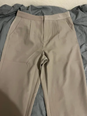 Beige kostymbyxor från 157, storlek M - Snygga beige kostymbyxor från 157 i storlek M. Byxorna har hög midja, raka ben och pressveck framtill. De är tillverkade i ett slätt material med diskreta fickor på sidorna och en bakficka. Stängs med dragkedja och knapp i midjan. Aldrig använda sen jag köpte de. 