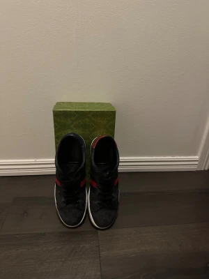 Gucci Ace - Snygga svarta Gucci sneakers med klassiskt GG-mönster, röd och blå rand på sidan samt orm skin-detaljer i rött och blått på hälen. Skorna har vit platt sula och svarta skosnören. Perfekta för dig som vill sticka ut i vinter. Allt og följer med förutom dustbag