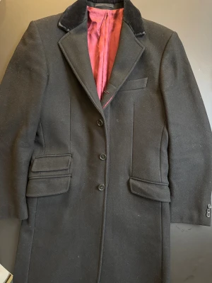 Svart rock från Merc London - Stilren svart rock (crombie coat) från Merc London med klassisk krage i sammet och snyggt vinrött innerfoder. Jackan har tre knappar framtill, flera fickor med lock och en elegant, rak passform. Perfekt för dig som vill ha en tidlös och sofistikerad look med inspiration från mods och teddy boys. Storleken passar mig perfekt (170 cm, 70 kg).