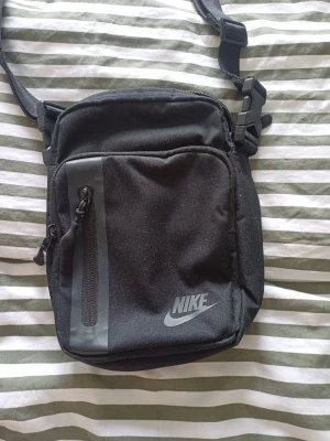 Svart axelväska från Nike - Snygg svart axelväska från Nike med justerbar rem och flera fack med dragkedja. Väskan har en stilren design med Nike-logga framtill och detaljer i grått. Perfekt för att bära med sig det viktigaste i vardagen.