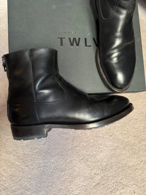Svarta boots från Project TWLV - Snygga svarta boots i Cordovan-skinn från Project TWLV med robust sula och dragkedja baktill. Klassisk rund tå, Blake Rapid-söm och stilren design som passar till många outfits. Skorna levereras i originalkartong med dustbag. Observera att angiven storlek från tillverkare är 41, Project TWLV:s märkning är alltid en storlek mindre än standard EU-storlek. Dessa sitter som stl 42 i andra märken, exempelvis RM Williams. Nypris ca 5000 kr.