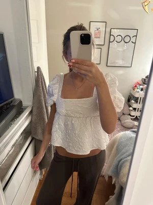 Vit blus med puffärm och rosett - Superfin vit blus med puffärmar och fyrkantig ringning. Blusen har ett broderat mönster i tyget och en söt rosett i ryggen. Croppad passform som ger en trendig look, perfekt till jeans eller kjol. Storlek xxs men passar upp till S.