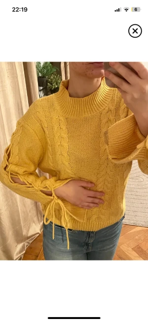 Gul stickad tröja med snörning - Snygg gul stickad tröja med grova flätstickade detaljer och cool snörning längs ärmarna. Tröjan har en hög, ribbad hals och långa ärmar med lite loose passform. Perfekt för dig som gillar att sticka ut med färg och detaljer.