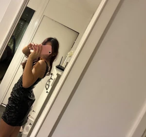 Svart glansig kort klänning - Säljer en svart glansig kort klänning med smala axelband och rynkade detaljer längs sidorna. Klänningen har en tight passform och är tillverkad i ett syntetmaterial som ger en cool, wet look-effekt. Perfekt för dig som vill sticka ut med en edgy stil.