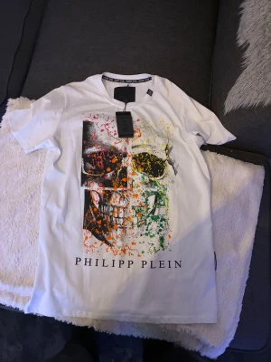 Vit t-shirt med döskalle från Philipp Plein - Snygg vit t-shirt från Philipp Plein i bomull med coolt döskalletryck framtill i svart, orange, gul, grön och rosa färg. Kortärmad modell med rund hals och logotypdetalj i nacken. Perfekt för dig som gillar statement-plagg och streetstyle.