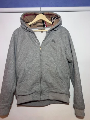 Grå Burberry hoodie med dragkedja - Grå hoodie från Burberry med klassiskt rutigt foder i huvan. Tröjan har dragkedja framtill, två fickor och broderad logga på bröstet. Tillverkad i mjuk bomull med ribbade muddar och huva med snörning. Perfekt för en chill och stilren look.
