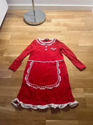 Röd julklänning med spets och volang - Supergullig röd långklänning med vita spetsdetaljer, volangkant nertill och söt ficka med polkagrisar. Klänningen har långa ärmar och en dekorativ vit krage samt ett stort förkläde framtill. Perfekt för julens alla tillfällen!