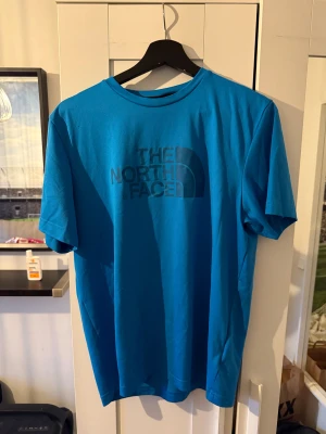 Blå t-shirt från The North Face - Snygg blå t-shirt från The North Face med stor logga tryckt på bröstet och mindre logga på ryggen. Klassisk rund hals och korta ärmar. Tillverkad i mjuk bomull som känns skön mot huden. Perfekt för dig som gillar enkel och stilren streetwear.