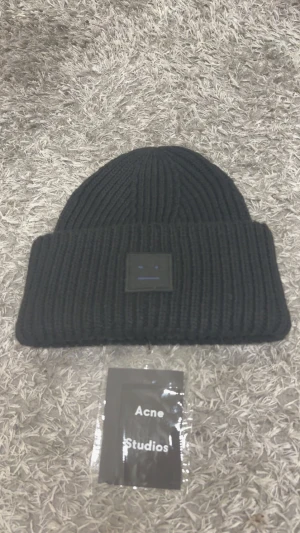 Acne Studios mössa - Svart ribbstickad mössa från Acne Studios med klassisk uppvikt kant och den ikoniska ansiktsloggan framtill. Tillverkad i mjukt ullmaterial som håller dig varm och snygg. Perfekt accessoar för kyliga dagar och enkel att matcha med din stil.