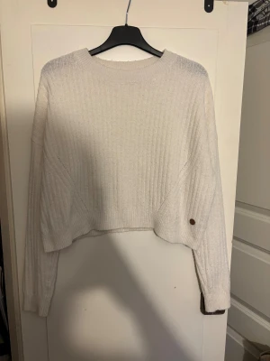 Vit ribbstickad croppad tröja Ü - Säljer en vit ribbstickad croppad tröja från Ü med rund hals och långa ärmar. Tröjan har en relaxed fit och en liten logga på ena sidan. Perfekt för lager-på-lager och chill dagar.