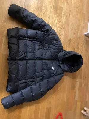 Svart pufferjacka från Nike Storm-FIT - Svart pufferjacka från Nike Storm-FIT med huva och quiltad design. Jackan har dragkedja med Nike-logga, två sidofickor och är tillverkad i syntetmaterial och dun, perfekt för kalla dagar och har en clean, sportig look.